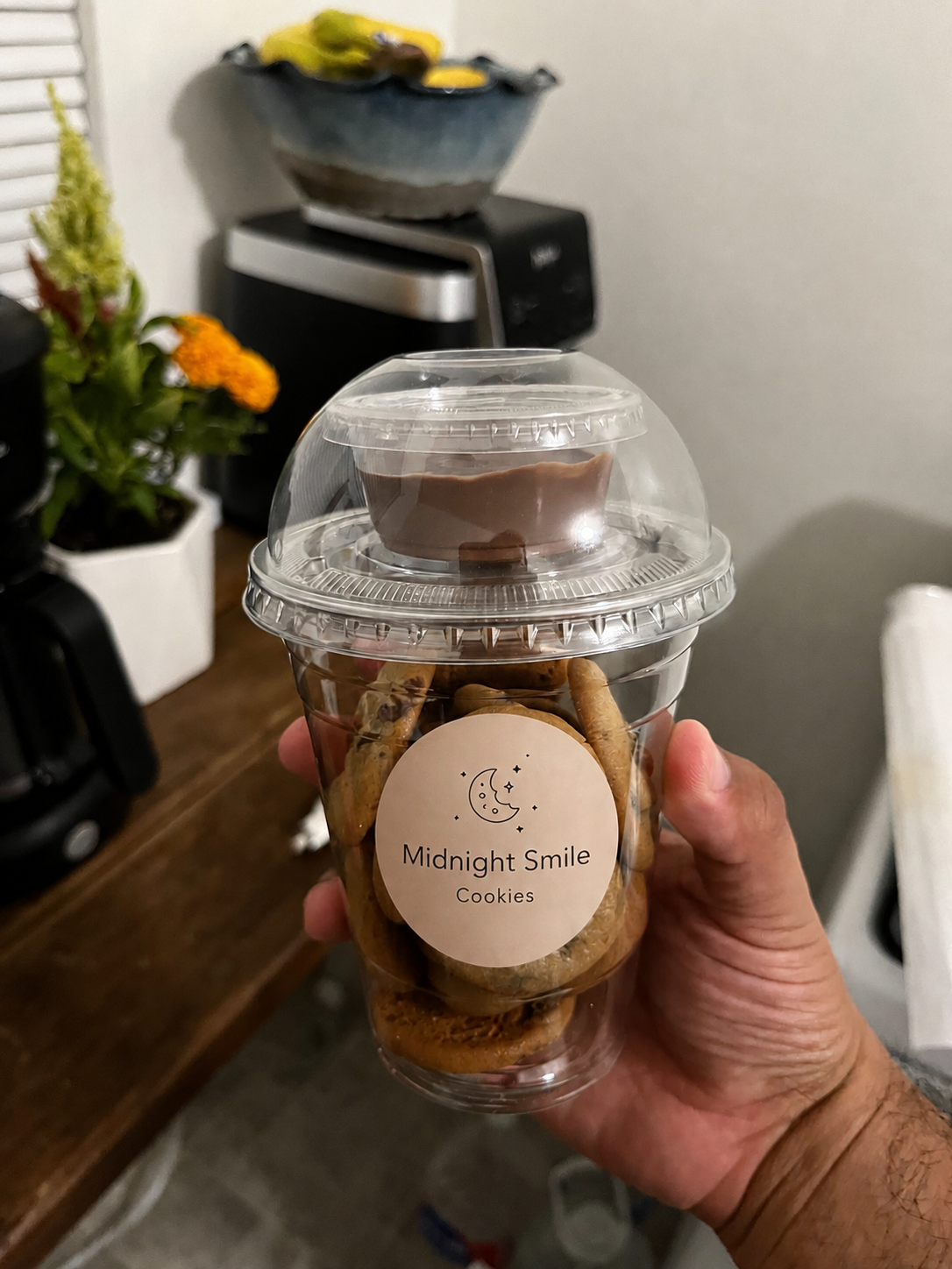 Hand holding a real Midnight Smile Cookies cup filled with mini chocolate chip cookies, topped with a domed lid and a cup of Nutella sauce